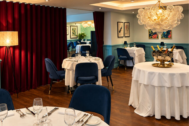 Elegant dining room at Chapter One Restaurant featuring white tablecloths, blue velvet chairs, and a glass chandelier.