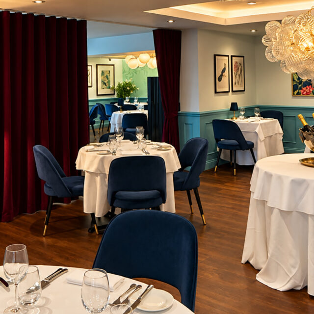 Elegant dining room at Chapter One Restaurant featuring white tablecloths, blue velvet chairs, and a glass chandelier.